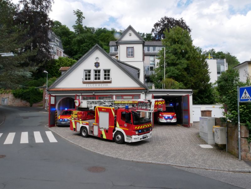 Feuerwehrhaus mit zwei geparkten Einsatzfahrzeugen in den Garagen und einem Löschfahrzeug vor dem Gebäude auf gepflastertem Hof