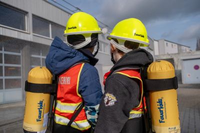 Zwei Kinder von hinten mit gelben Feuerwehrhelmen und gelben Atemschutzflaschen auf dem Rücken, sie tragen rote Warnwesten