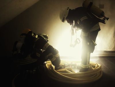 Zwei Feuerwehrleute in Schutzkleidung und Atemschutzgeräten rollen einen Schlauch in einem dunklen Raum mit starkem Licht im Hintergrund